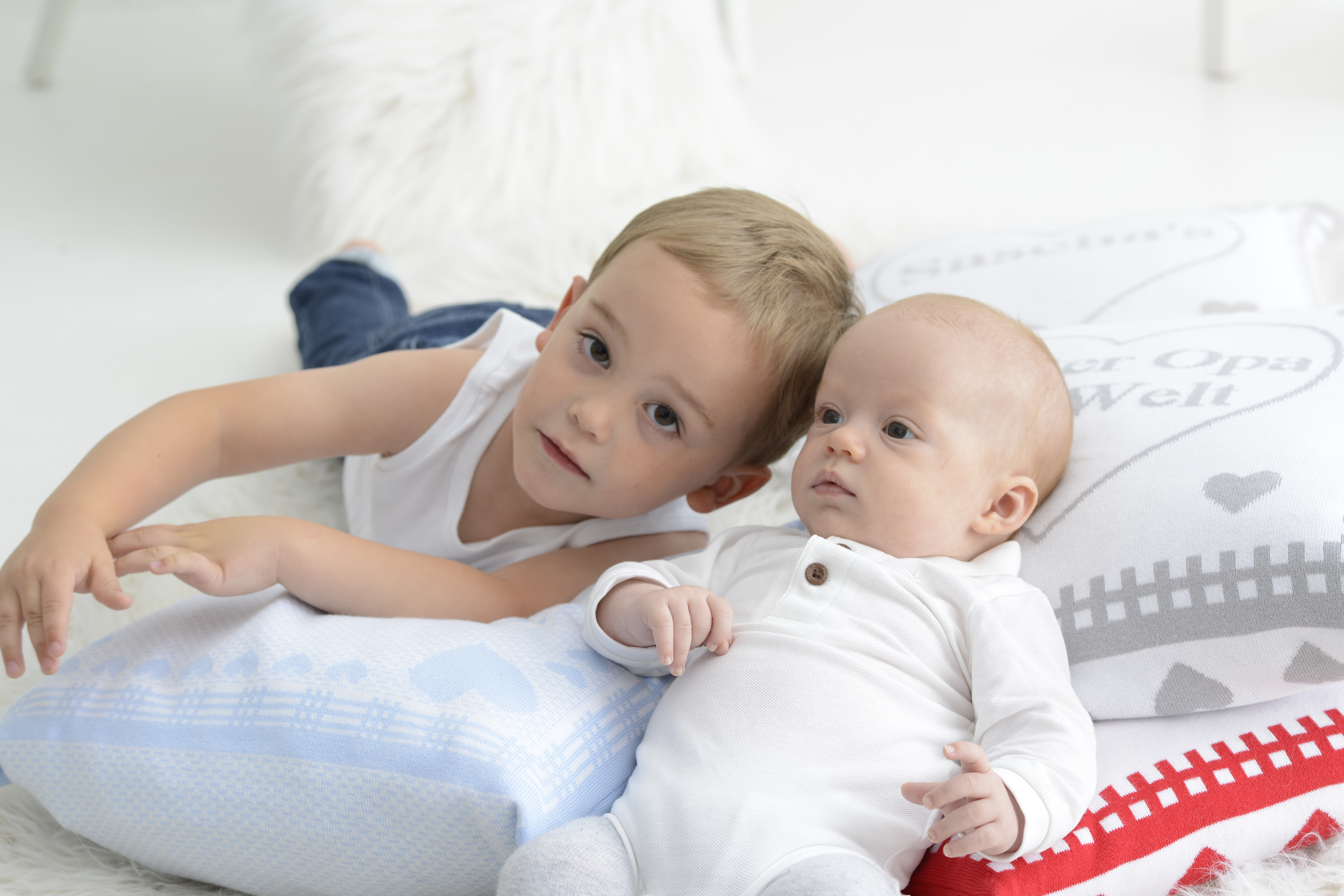 Blonder Junge schaut in die Kamera, neben kleinem Baby in weißem Strampelanzuh. Sie liegen auf personalisierten Kissen in blau und rot.